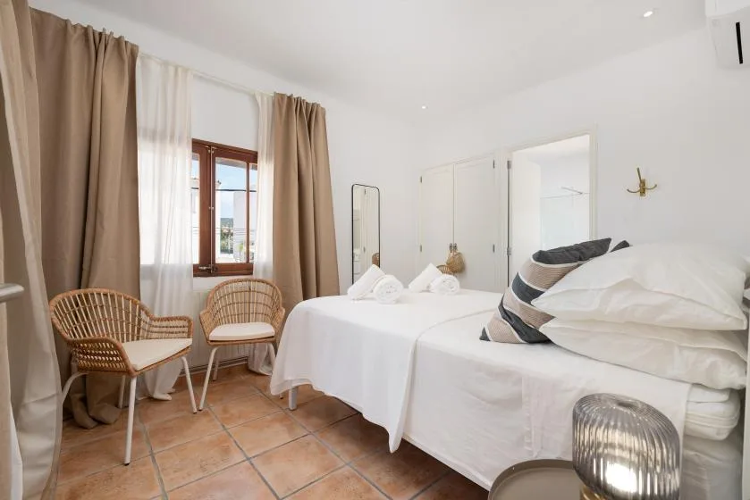 A bright bedroom with white bedding, tall curtained windows, and two seats.