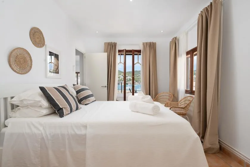 A bright bedroom with a neatly made bed, woven chairs, and tall windows.