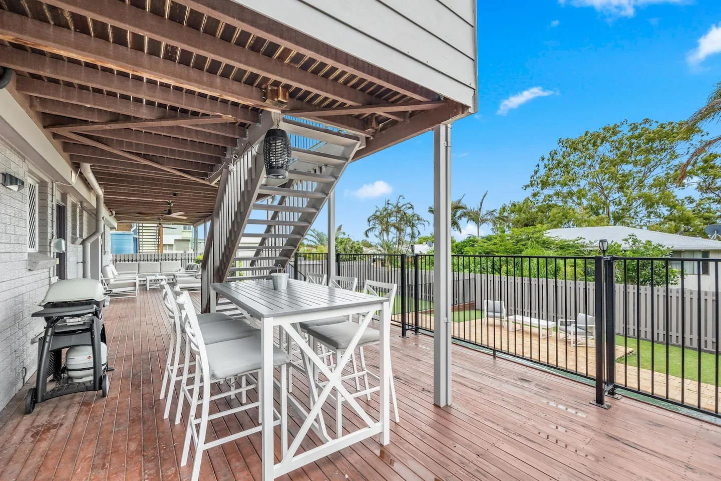 An outdoor dining area with a barbecue sits beneath a raised deck beside a fenced yard.