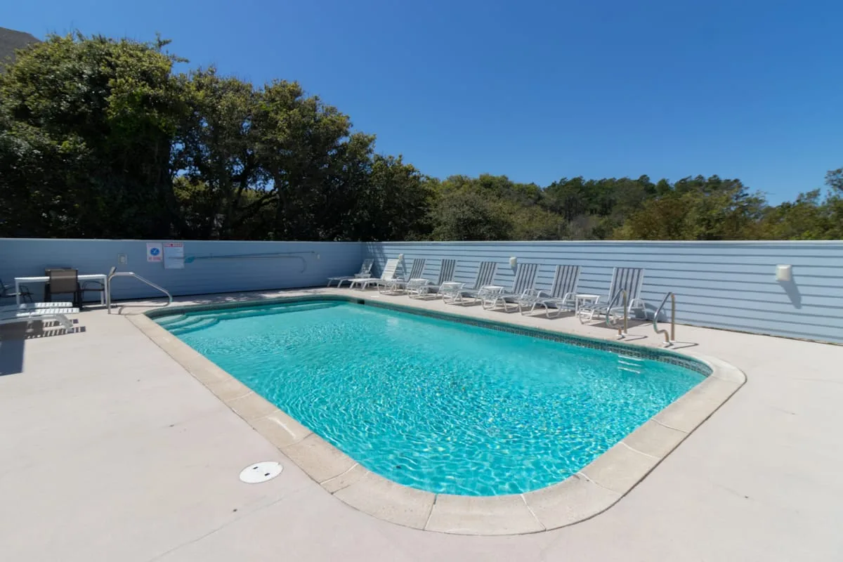 An outdoor swimming pool sits beside a row of lounge chairs under a clear sky.