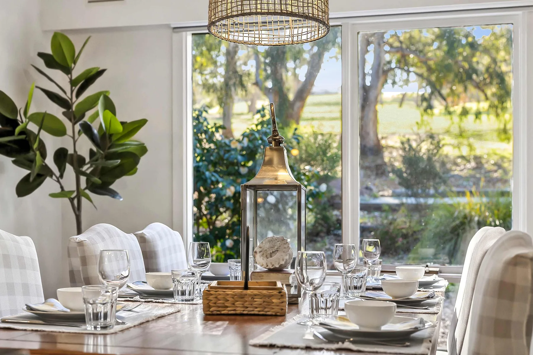 A dining table is set with plates and glassware beside large windows.