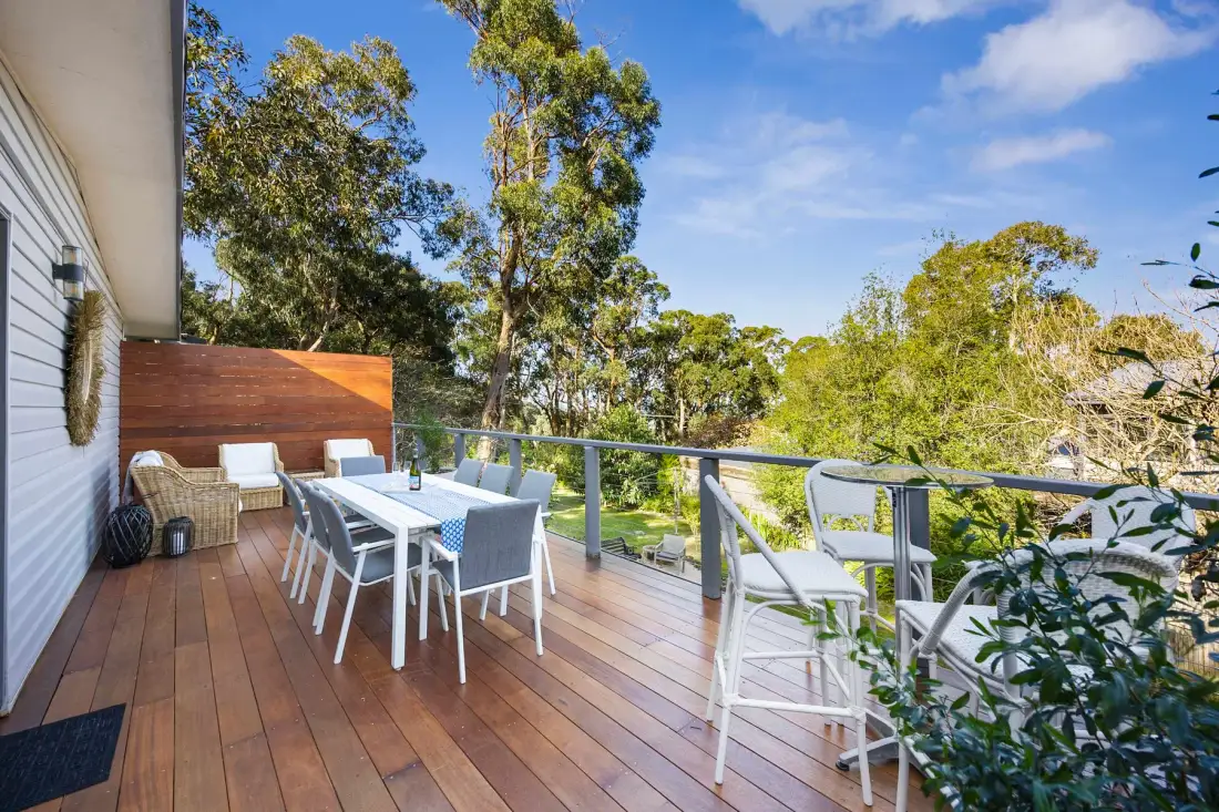 A wooden outdoor deck has a dining table and seating overlooking leafy trees.