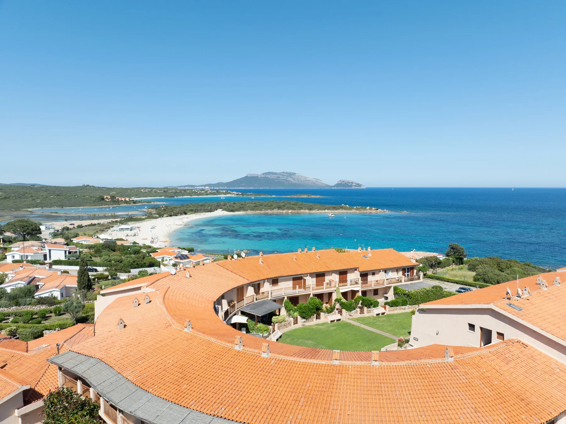 Vista aérea de un complejo costero con tejados de terracota junto a una bahía turquesa.