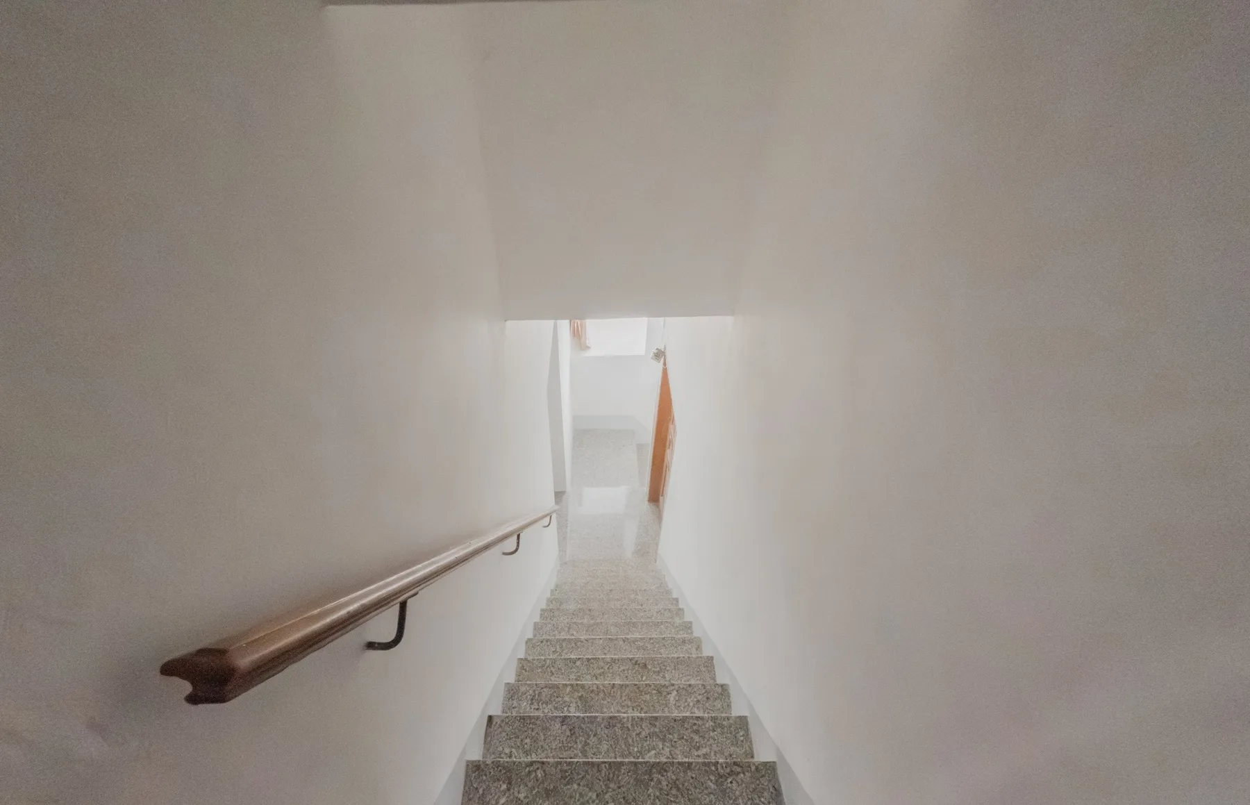 A narrow indoor stairwell descends between white walls with a wooden handrail.