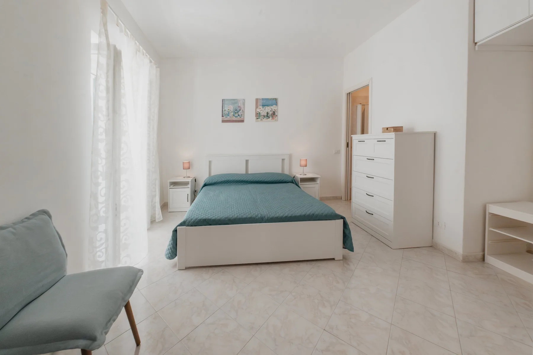 A bright bedroom with a teal-covered bed, bedside tables, and a white dresser.