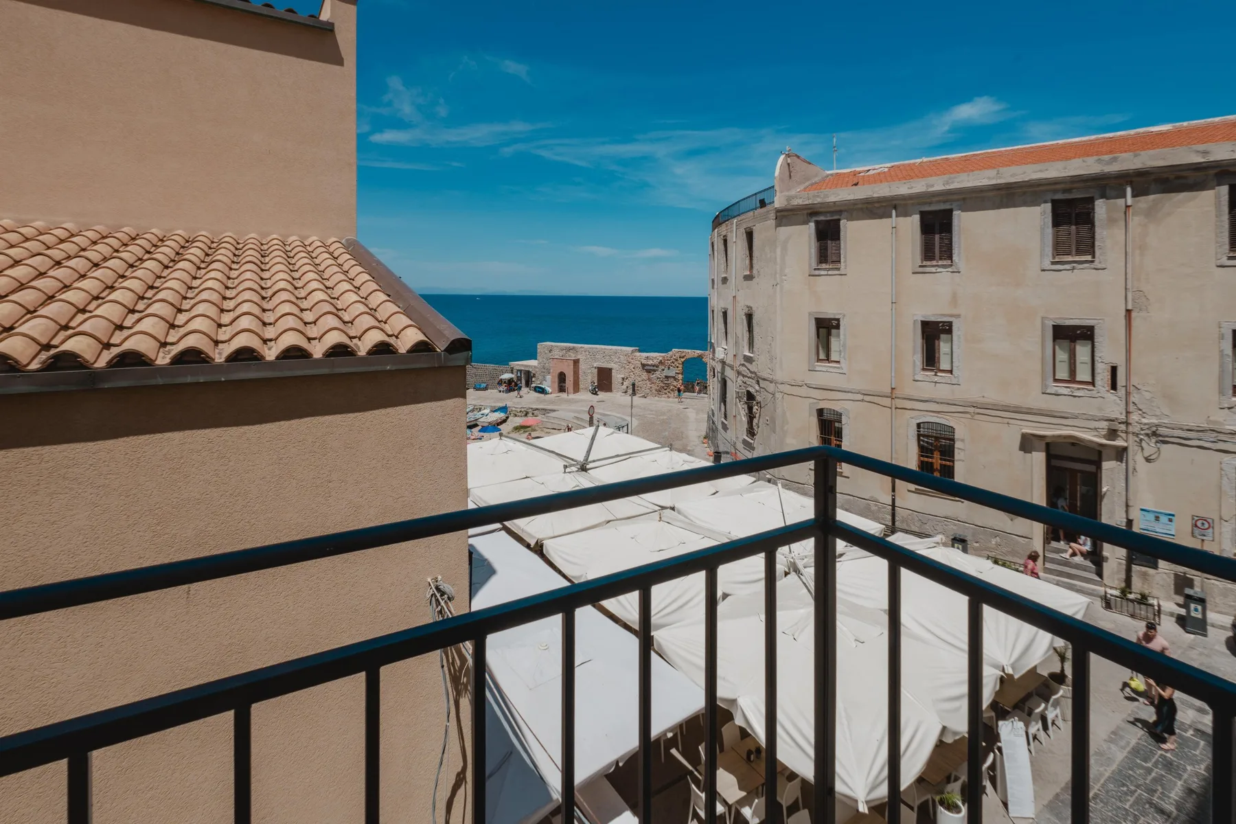 A balcony overlooks a seaside square with white canopies and surrounding buildings.