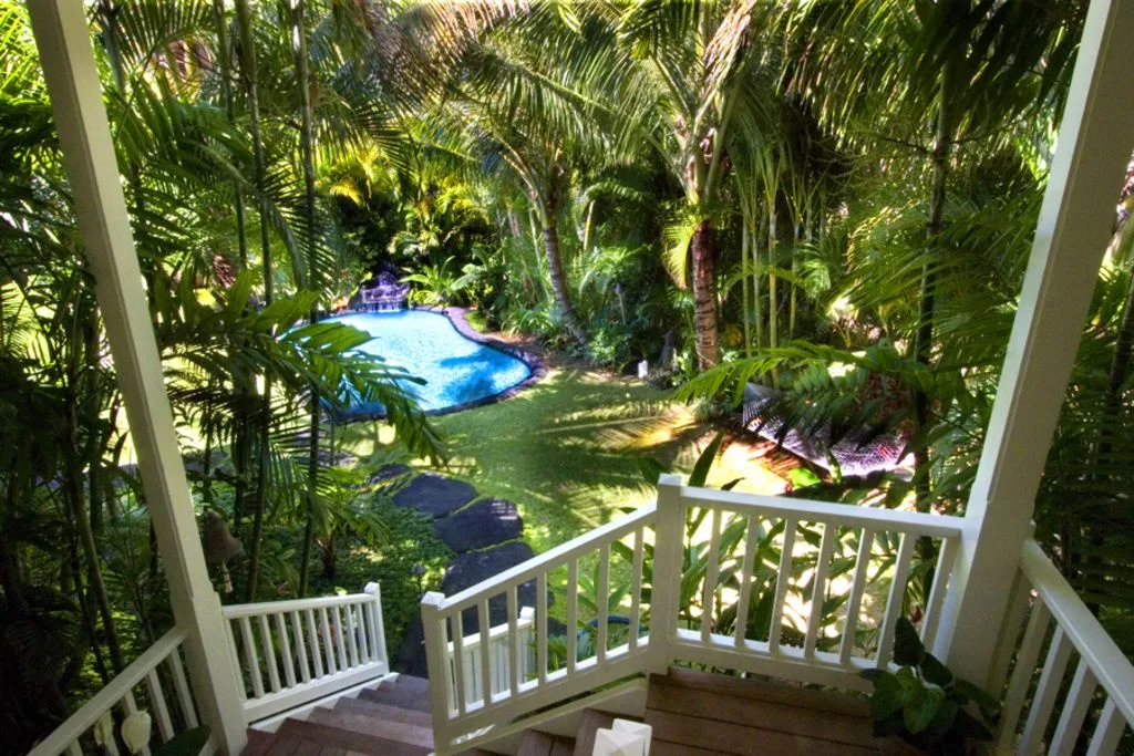 A leafy garden surrounds a blue swimming pool beside a wooden deck and white railing.