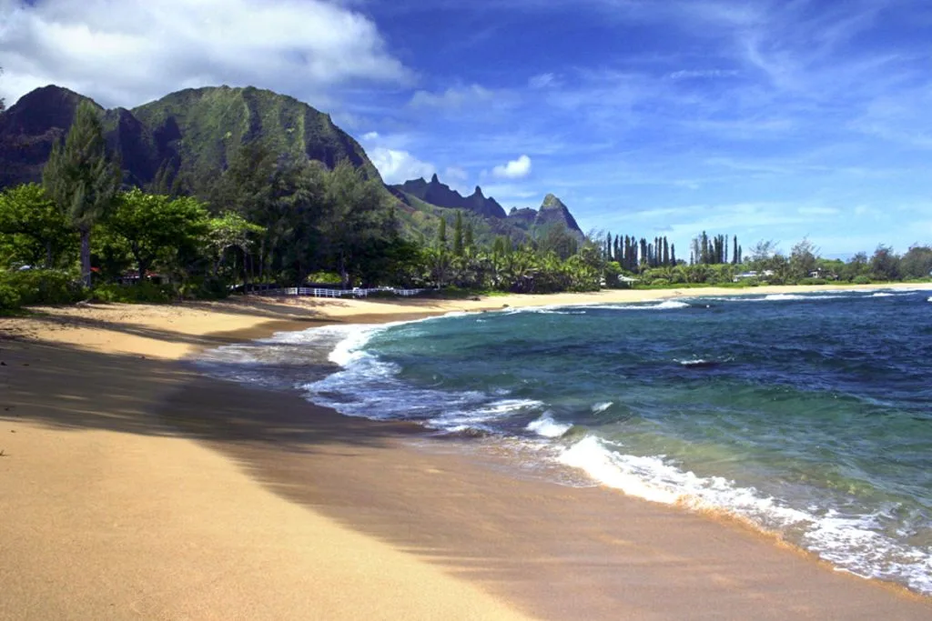 A sandy beach curves along blue water beneath green mountains.