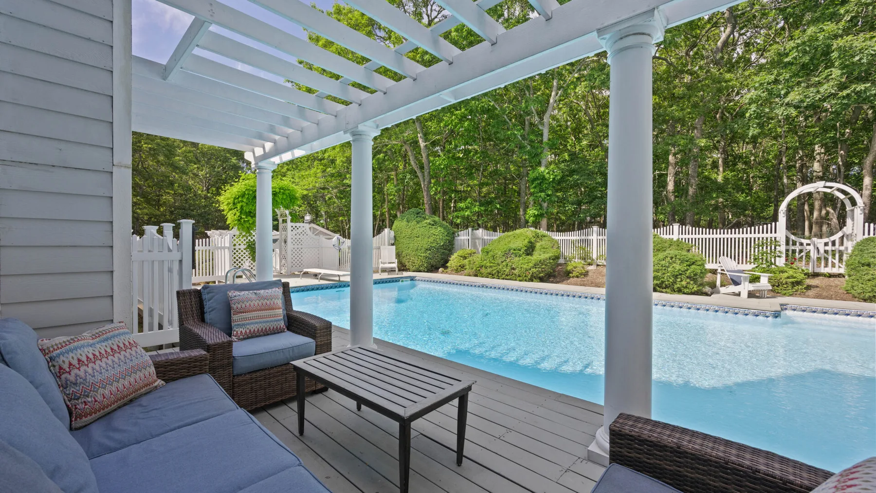 Una zona exterior cubierta con asientos da a una piscina y un jardín cercado.