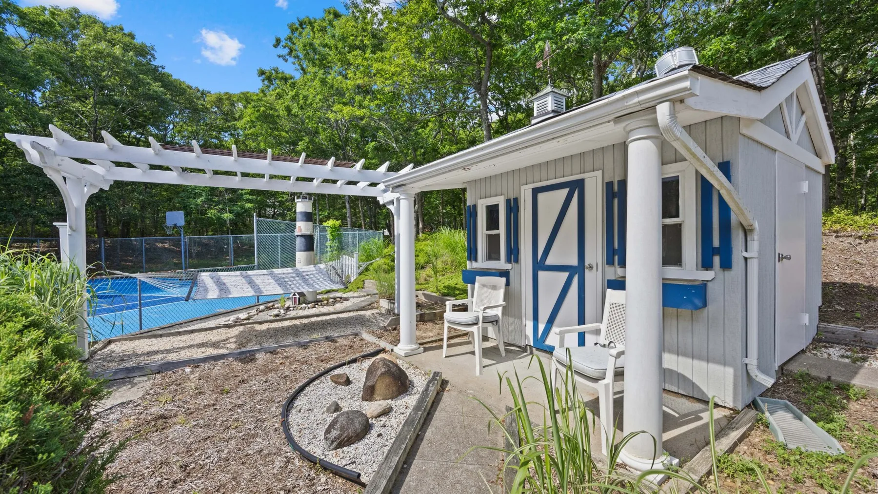 Una zona de piscina al aire libre junto a un pequeño edificio claro y una pérgola blanca.