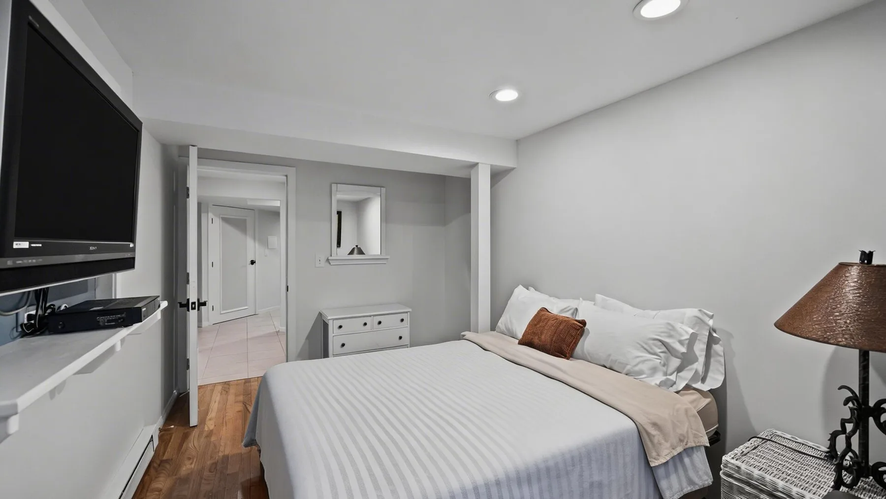Un dormitorio con una cama bien hecha, un televisor en la pared y una lámpara de noche.
