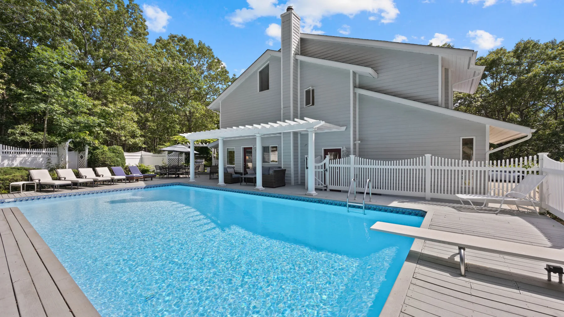Una piscina junto a una casa moderna gris, rodeada por una terraza de madera y una cerca blanca.