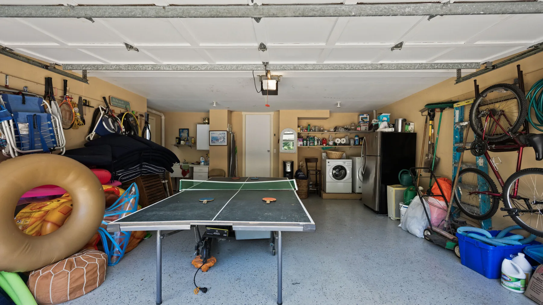 Un garaje con una mesa de ping-pong, estantes de almacenamiento y bicicletas junto a las paredes.