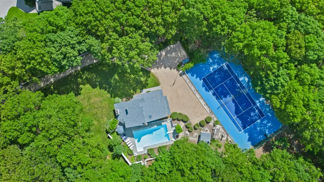 Vista aérea de una casa, una piscina y una cancha de tenis rodeadas de árboles.