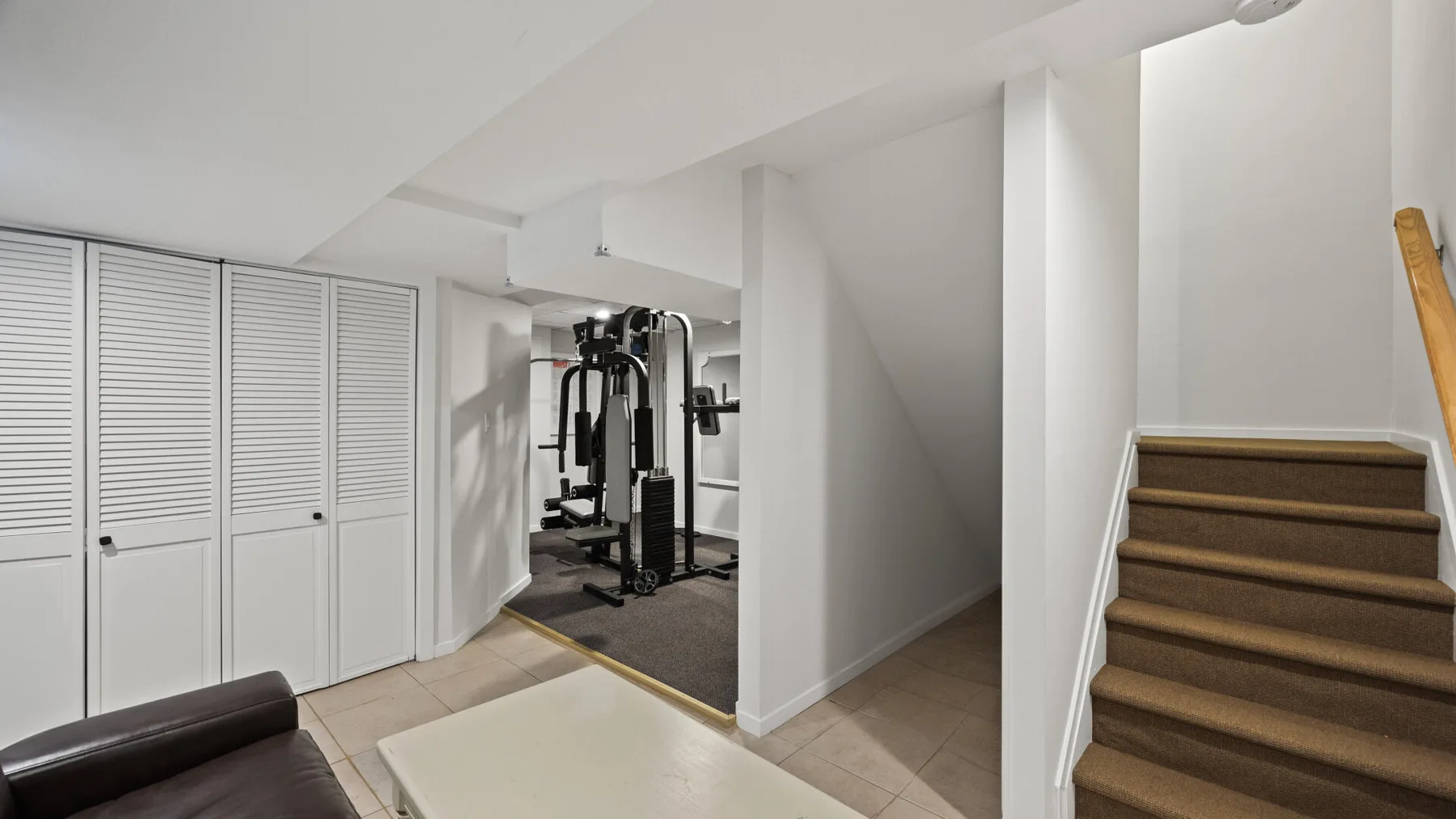 Un gimnasio doméstico con equipo de ejercicio está junto a una escalera y puertas de almacenamiento.