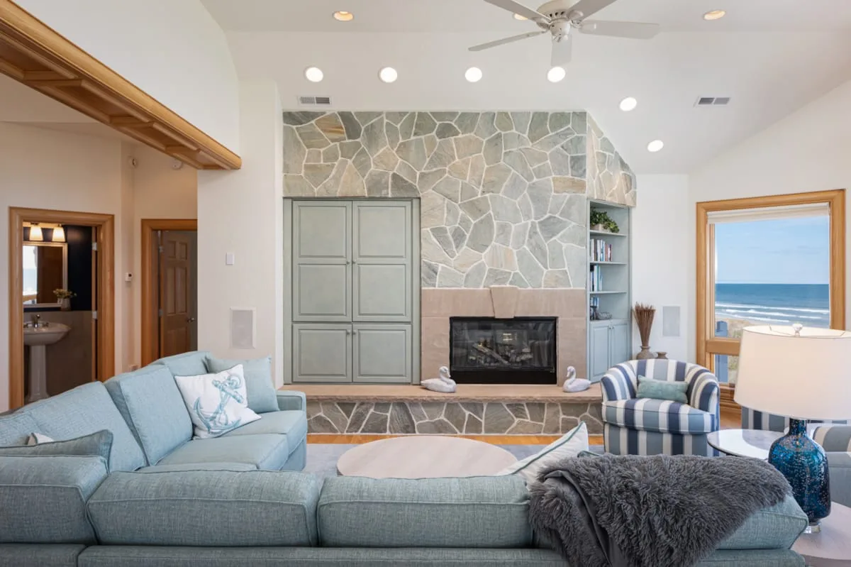 A living room with seating around a stone fireplace and a view of the ocean.