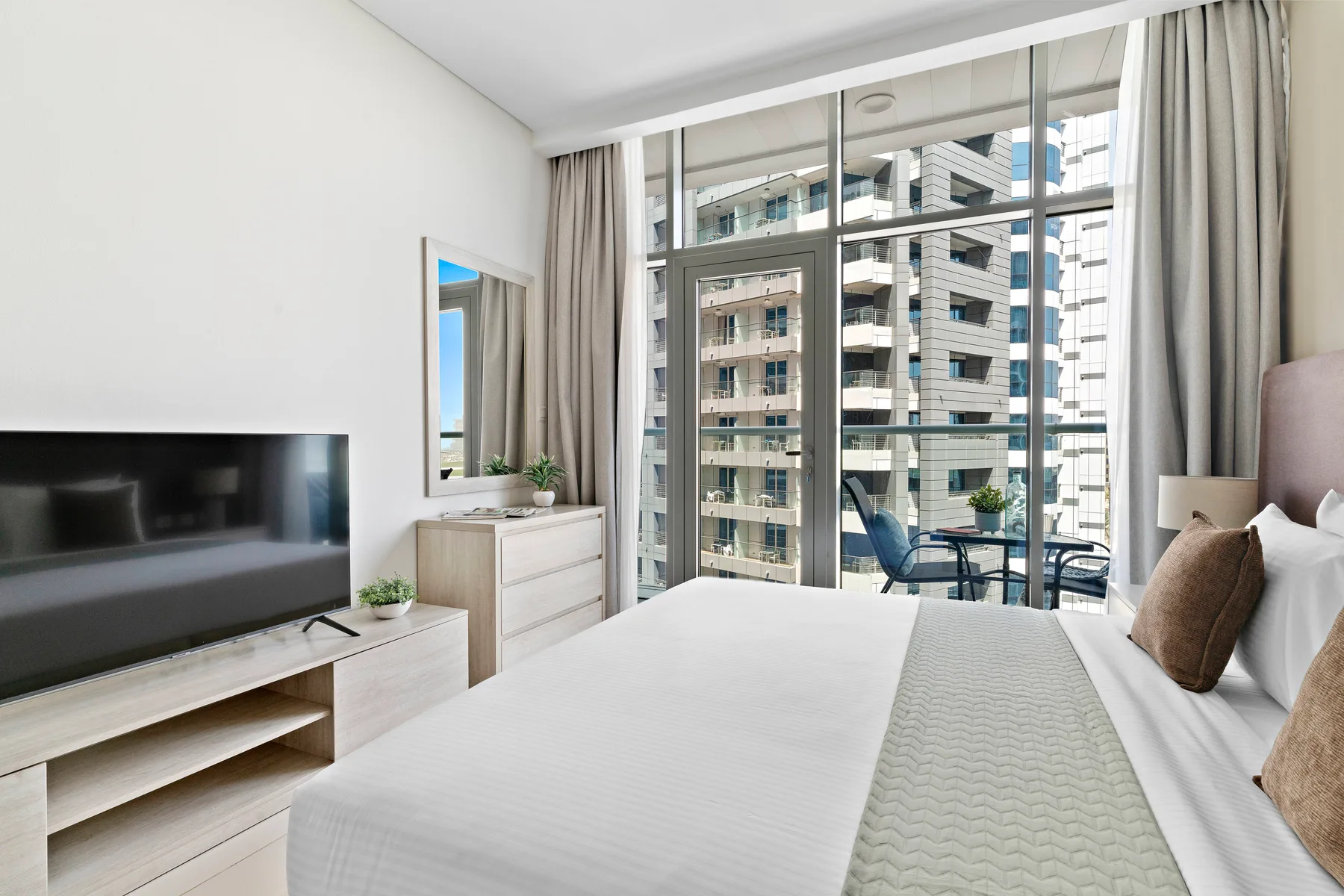 Un dormitorio luminoso con una cama tendida y grandes ventanas con vista a edificios cercanos.