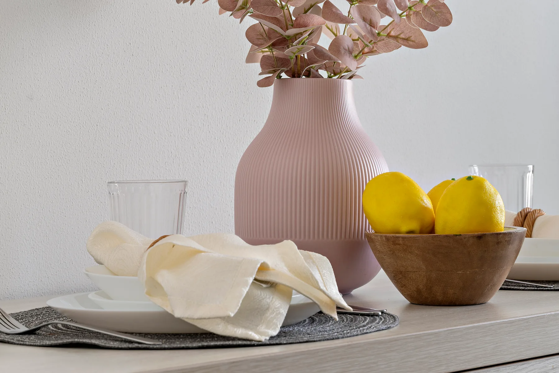 Una mesa de comedor está puesta con platos, limones y un jarrón con flores secas.
