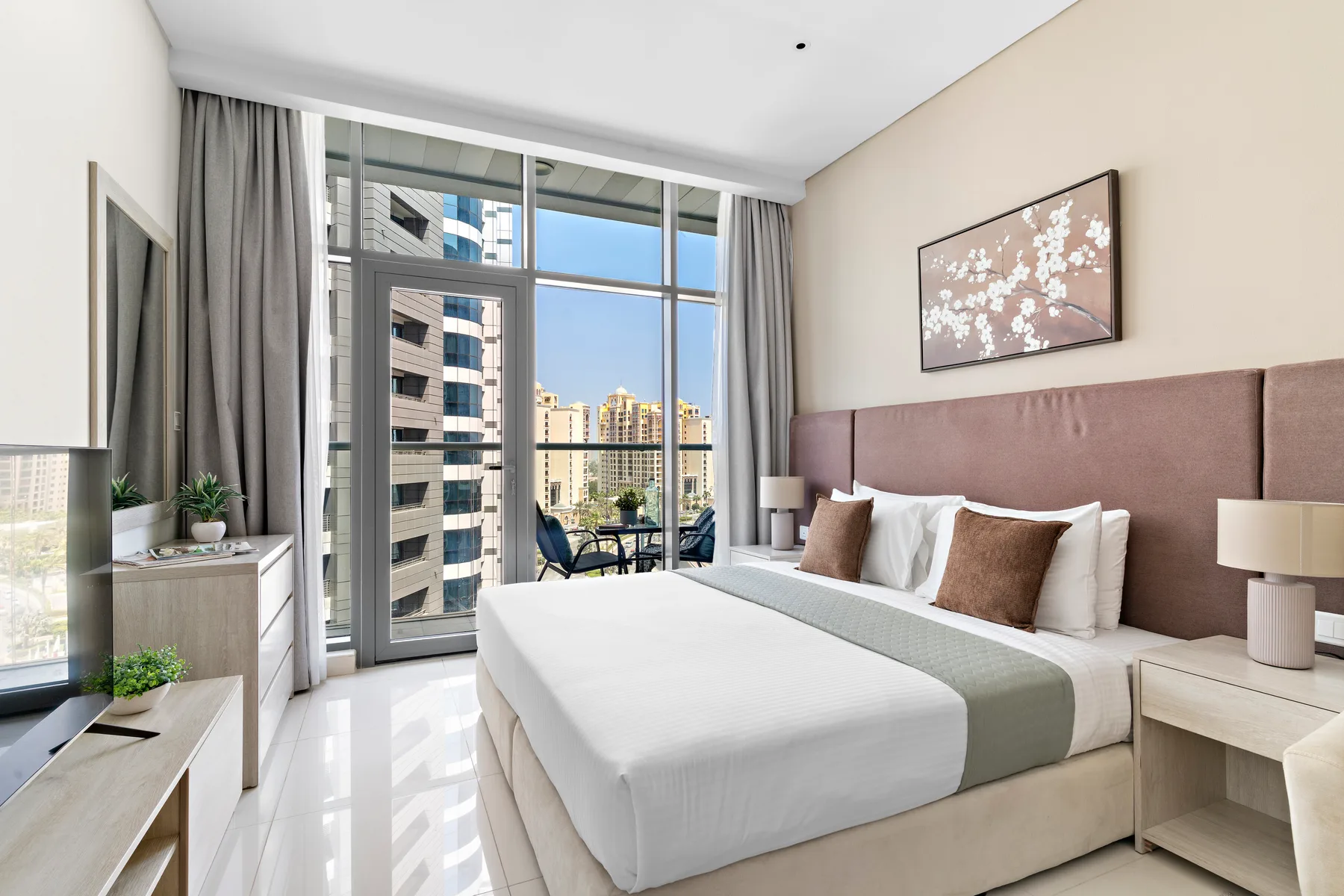A bedroom with a neatly made bed and large windows overlooking nearby buildings.
