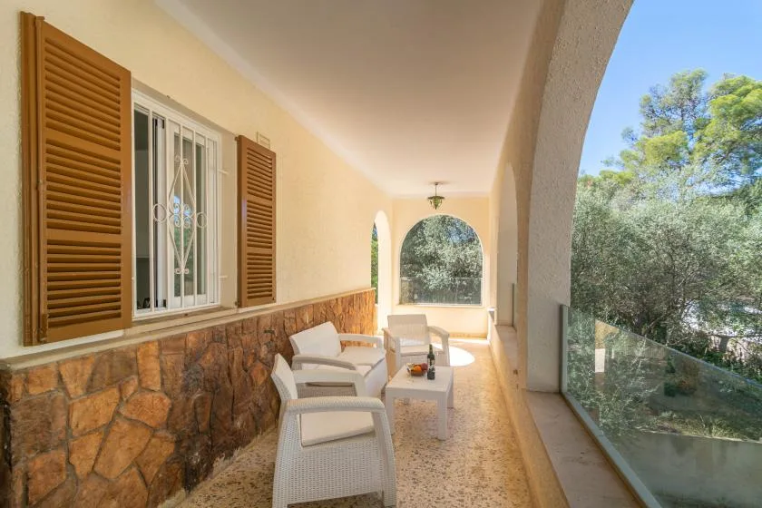 A covered outdoor seating area has pale furnishings beside shuttered windows.