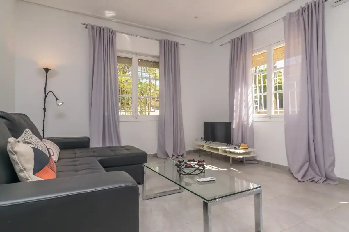 A bright living room with seating, a television, and tall curtained windows.