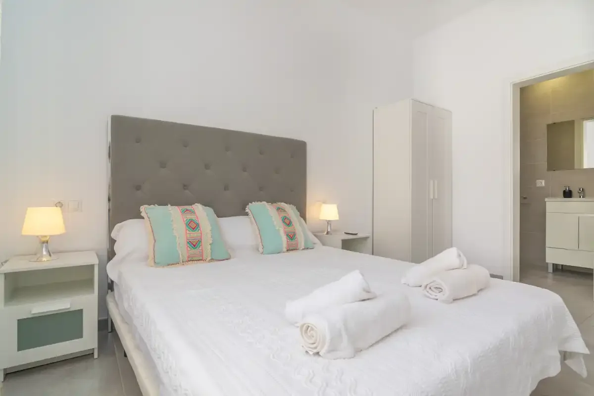A bright bedroom with a double bed, bedside tables, and an open bathroom doorway.
