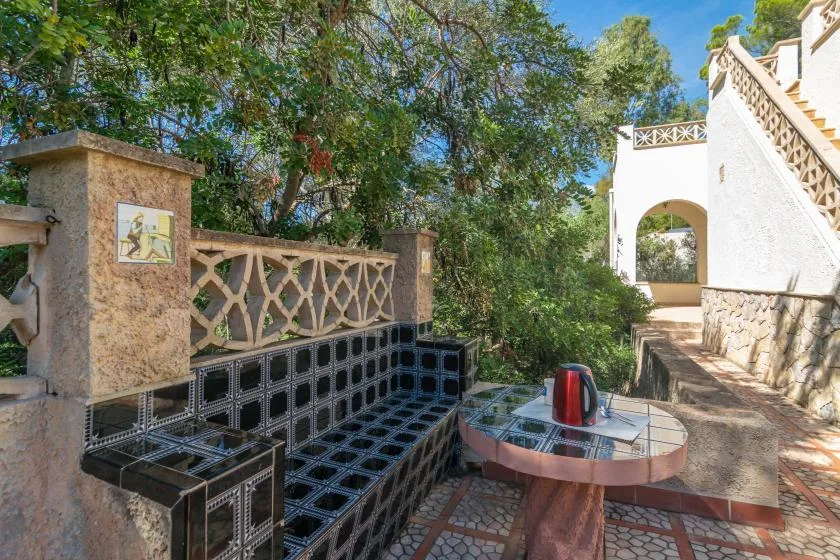 A tiled outdoor patio has built-in seating and a round table beneath leafy trees.