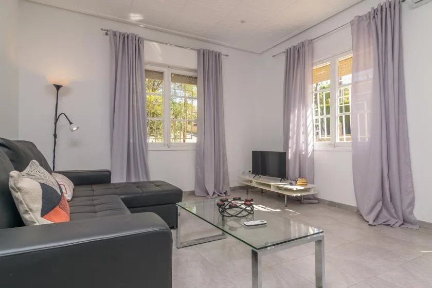 A bright living room with dark seating, a glass coffee table, and tall curtained windows.