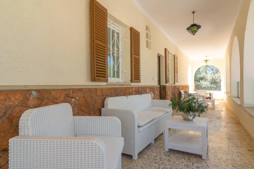 A covered patio has white seating, a small table, and an arched opening.