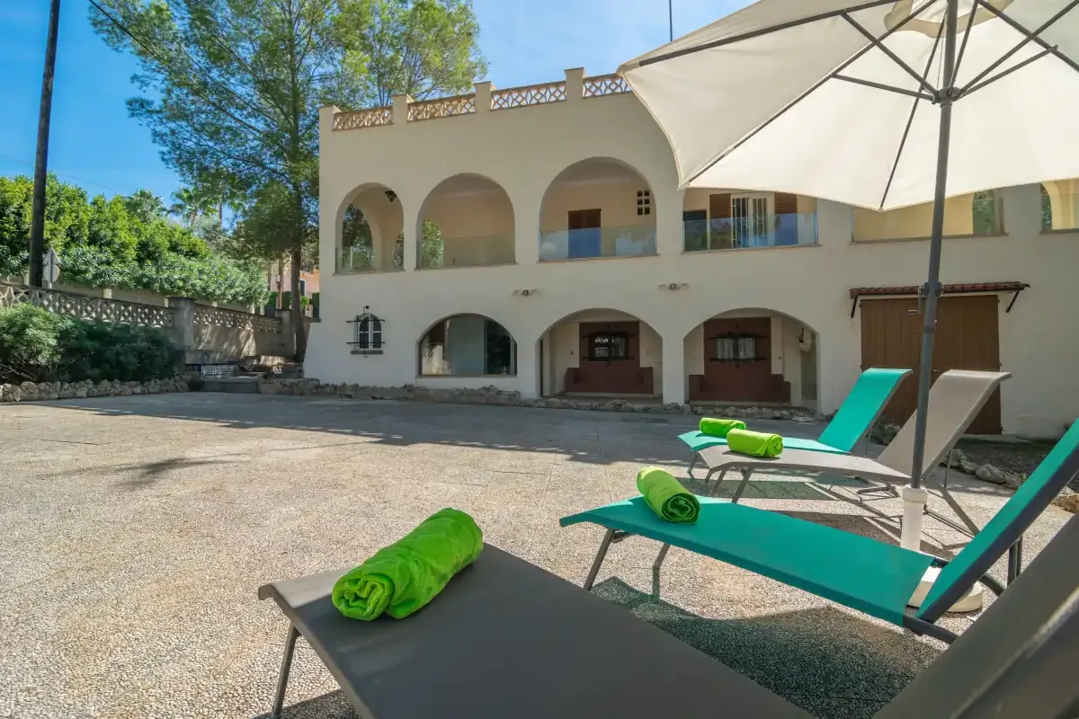 A sunny courtyard has turquoise loungers and a two-story building with arched openings.