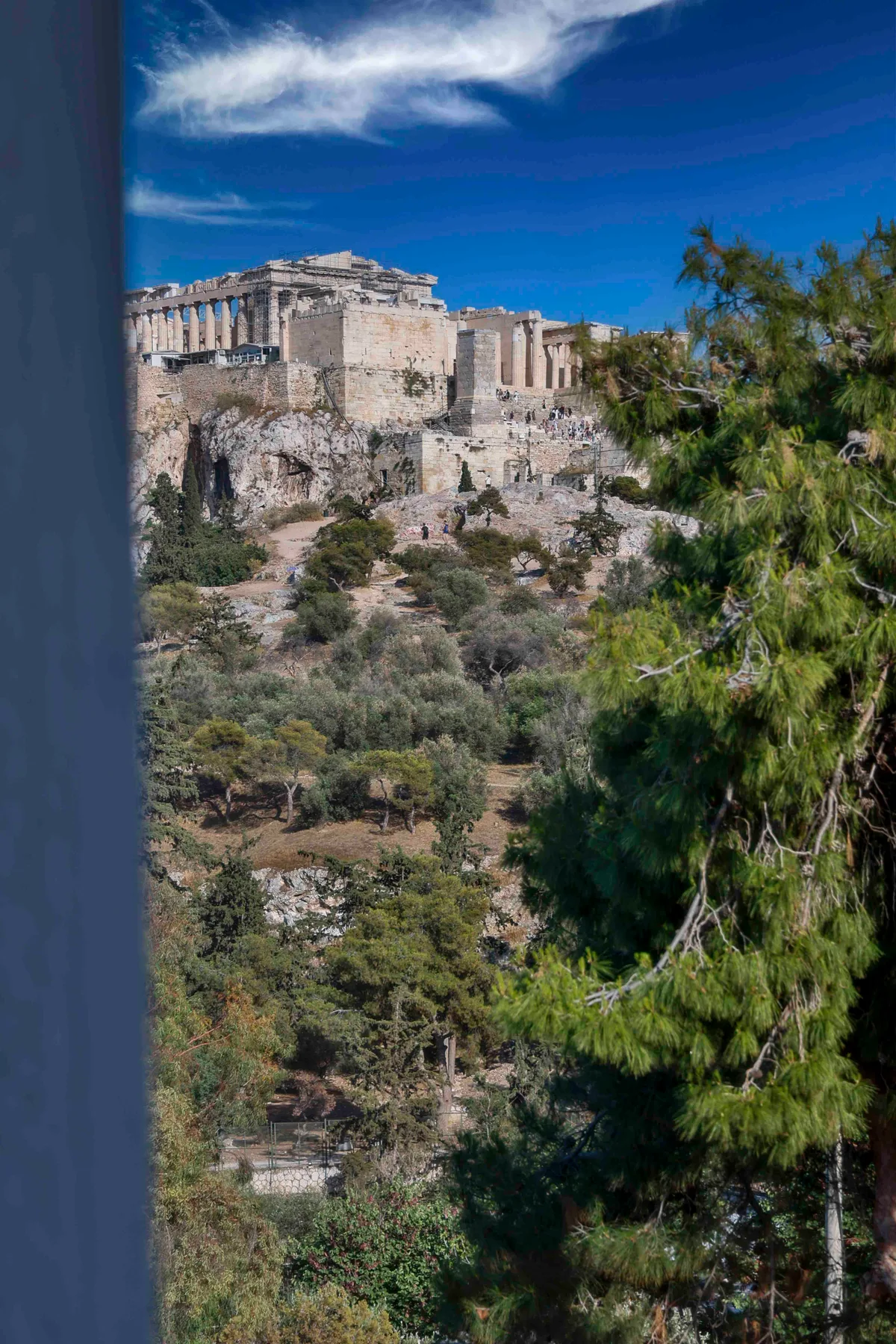 The Acropolis and Parthenon rise above a wooded hillside beneath a blue sky.