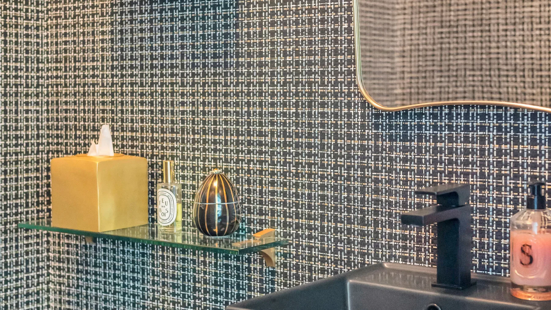 Un lavabo de baño con grifo oscuro, repisa de vidrio y pared estampada.