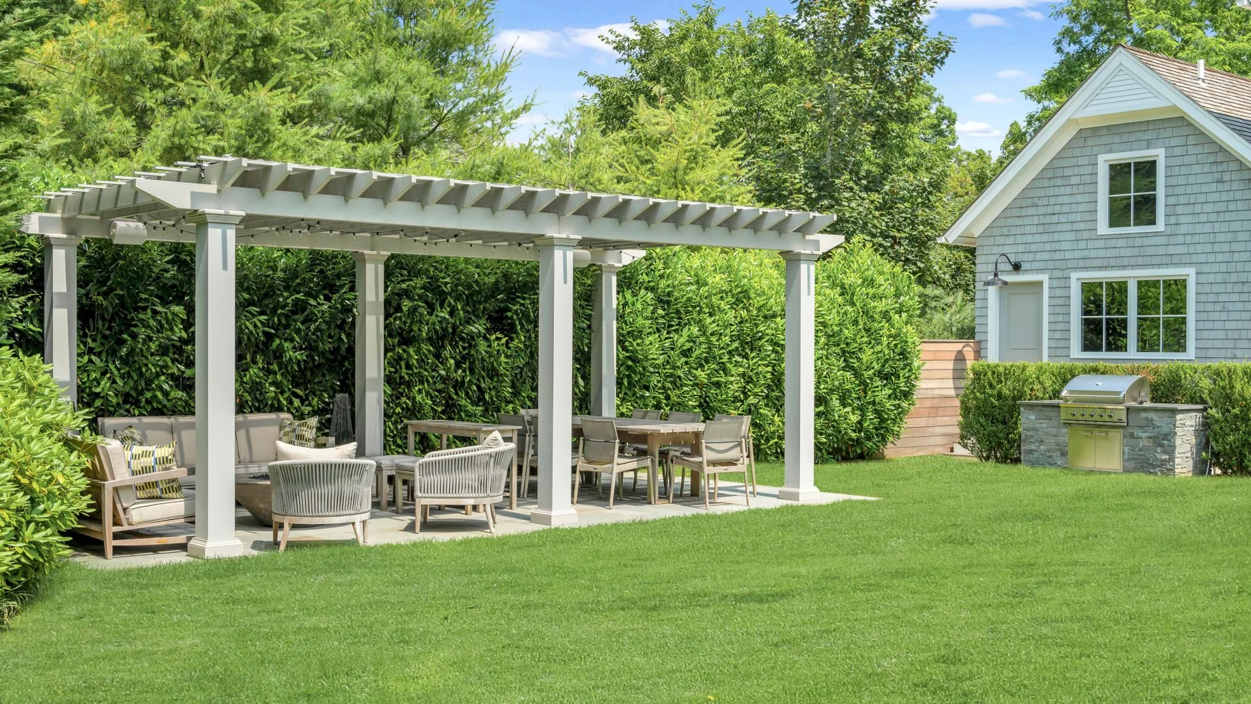 A garden patio with a pergola, outdoor seating, and a table on a green lawn.