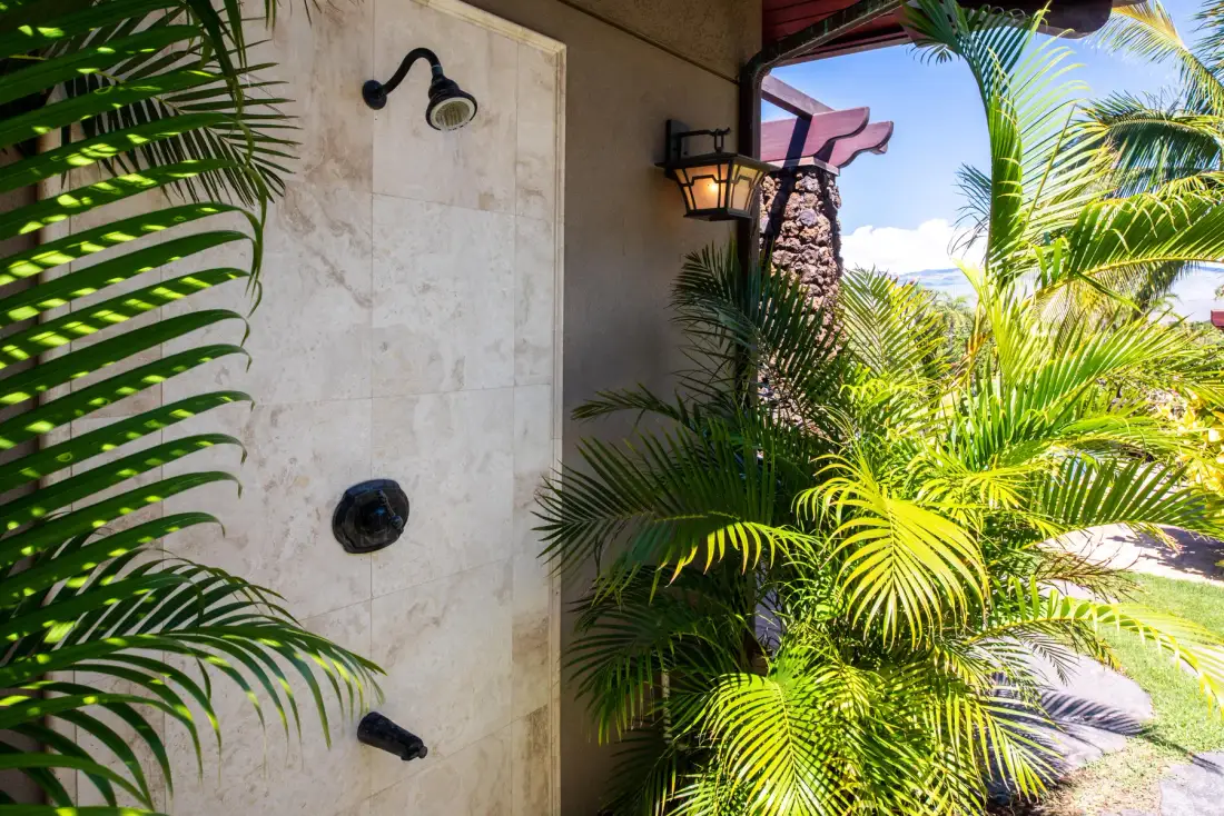 An outdoor shower beside a wall, surrounded by lush tropical plants.