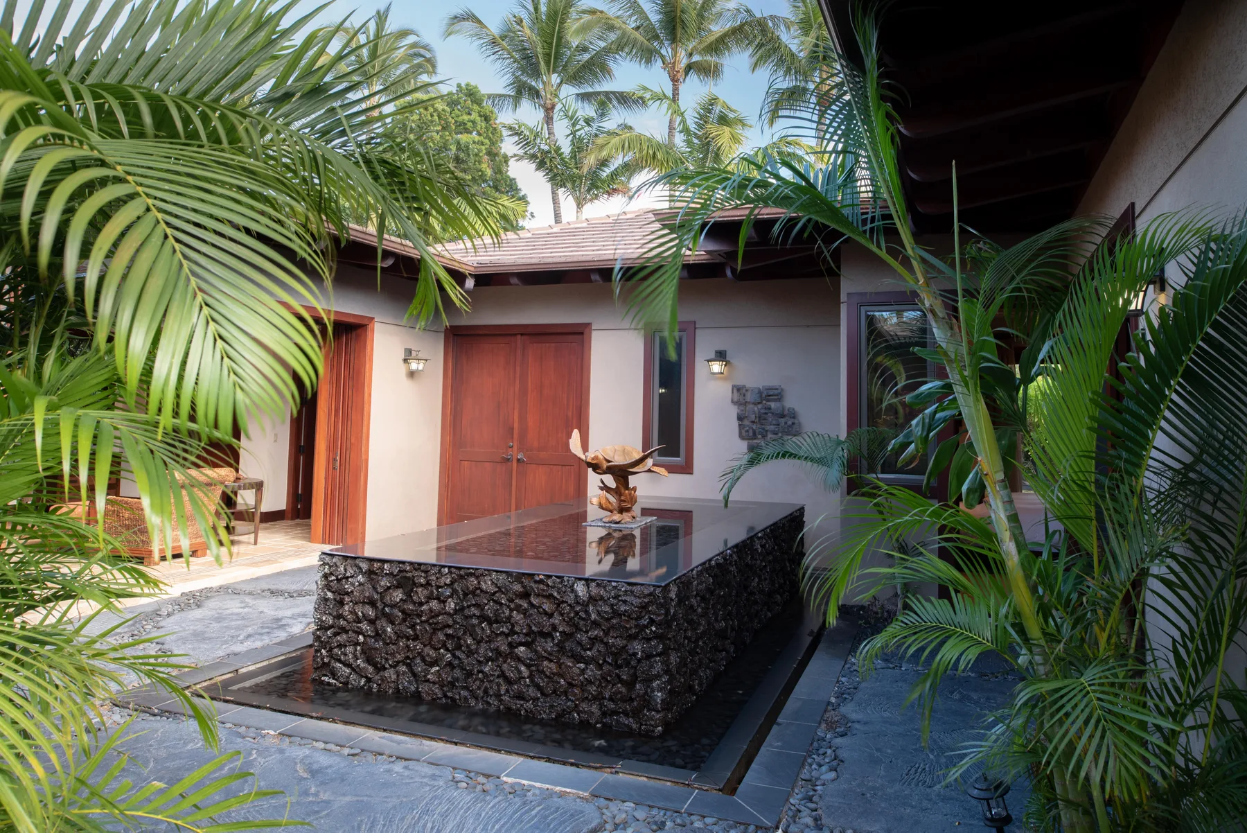 A tropical courtyard features a stone water basin surrounded by leafy plants.
