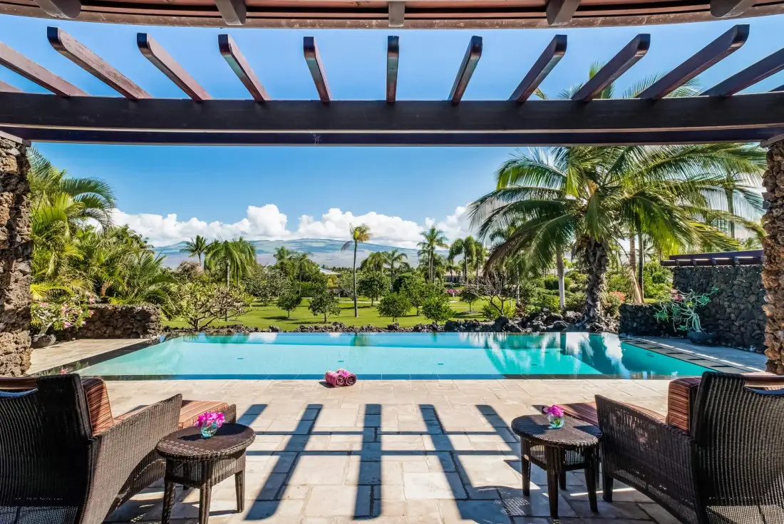 A swimming pool and paved patio overlook a tropical garden beneath a wooden pergola.