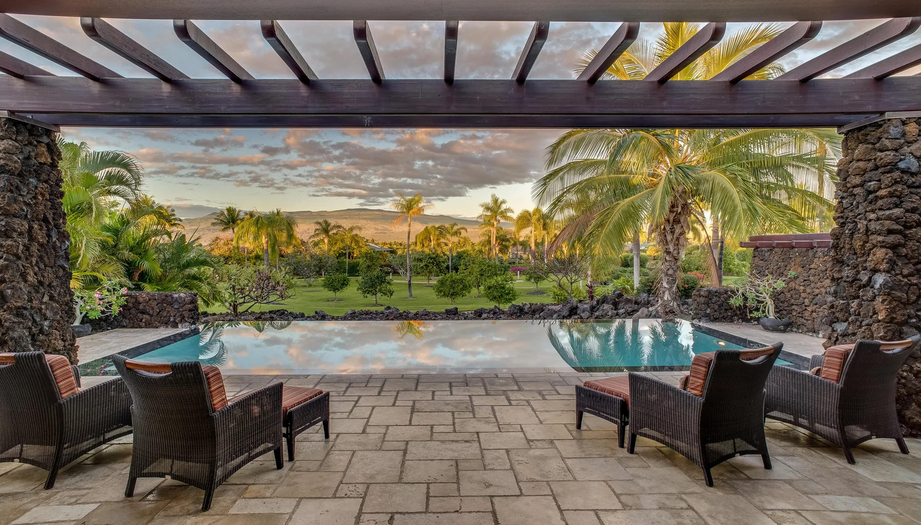 An outdoor pool and paved patio sit beneath a pergola, facing a palm-lined garden.