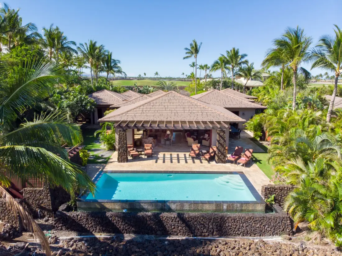 A tropical outdoor pool sits in front of a shaded pavilion surrounded by palm trees.