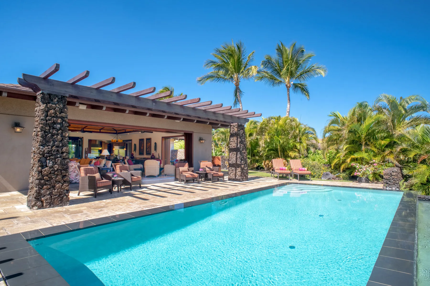 Outdoor pool beside a covered patio with seating and palm trees.