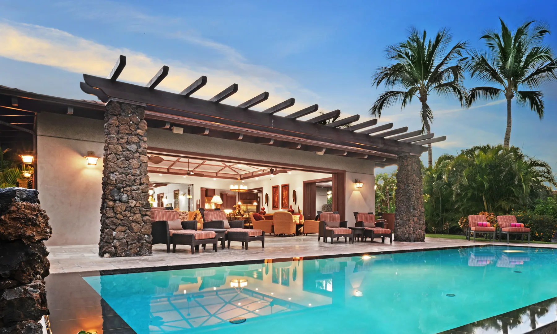 An outdoor swimming pool sits in front of an open-sided lounge with palm trees nearby.
