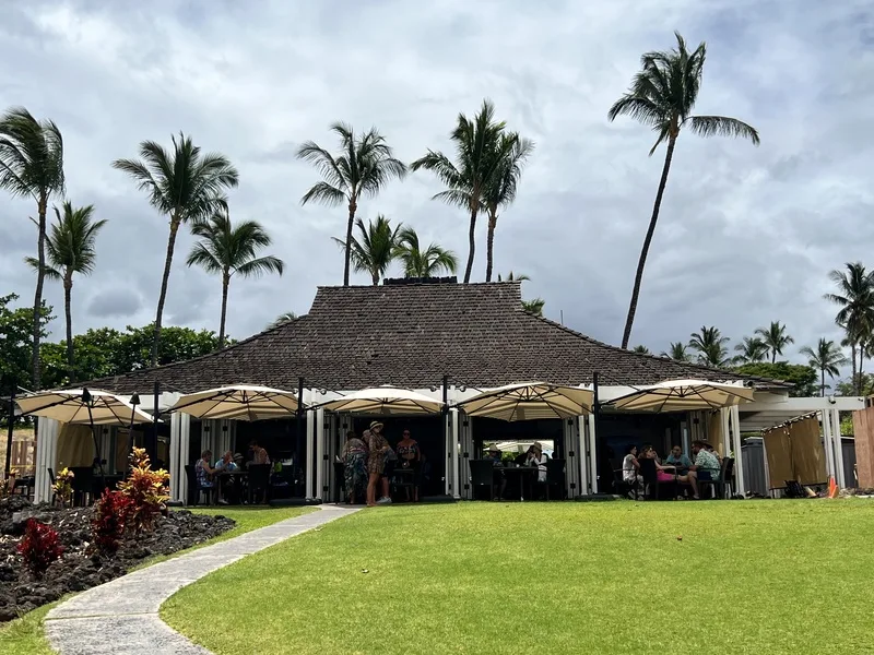 An open-air pavilion with a broad roof and patio umbrellas stands beside a lawn.