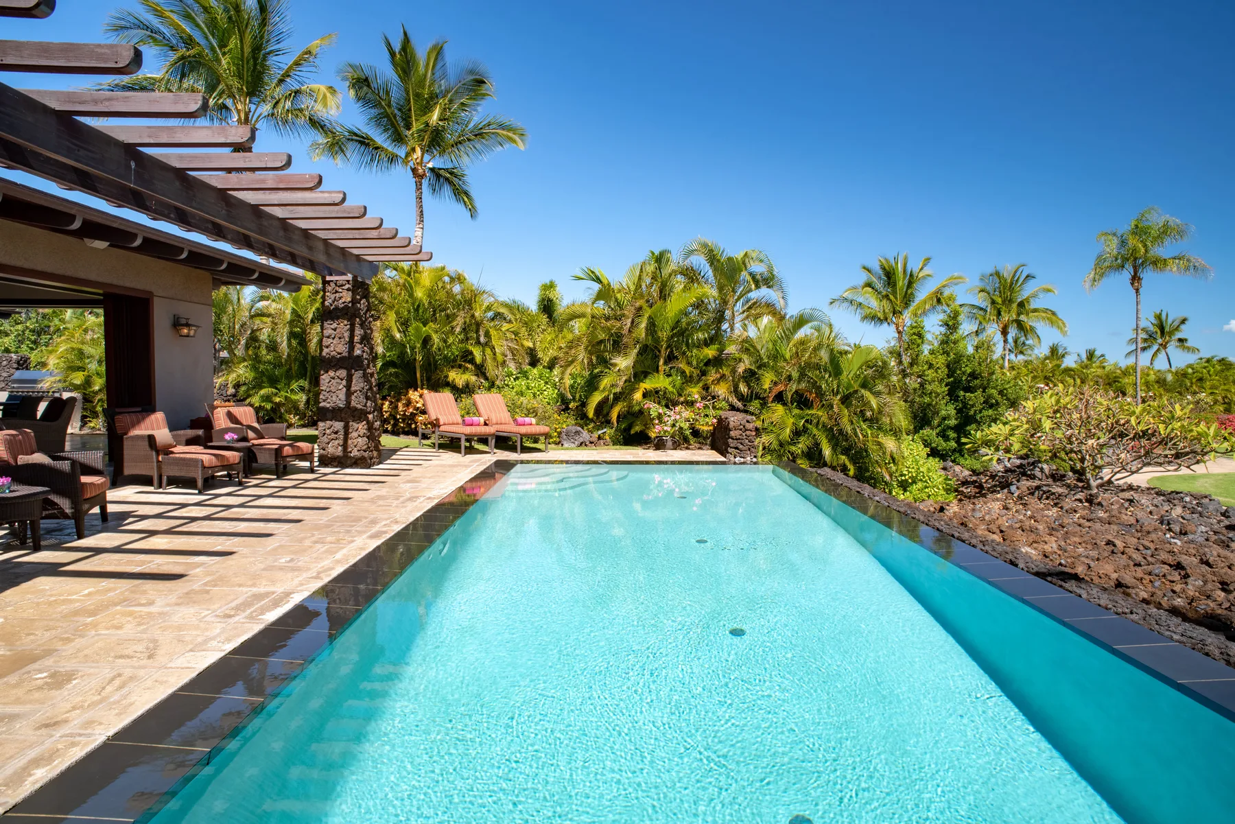 An outdoor swimming pool beside a tiled patio with seating and palm trees.