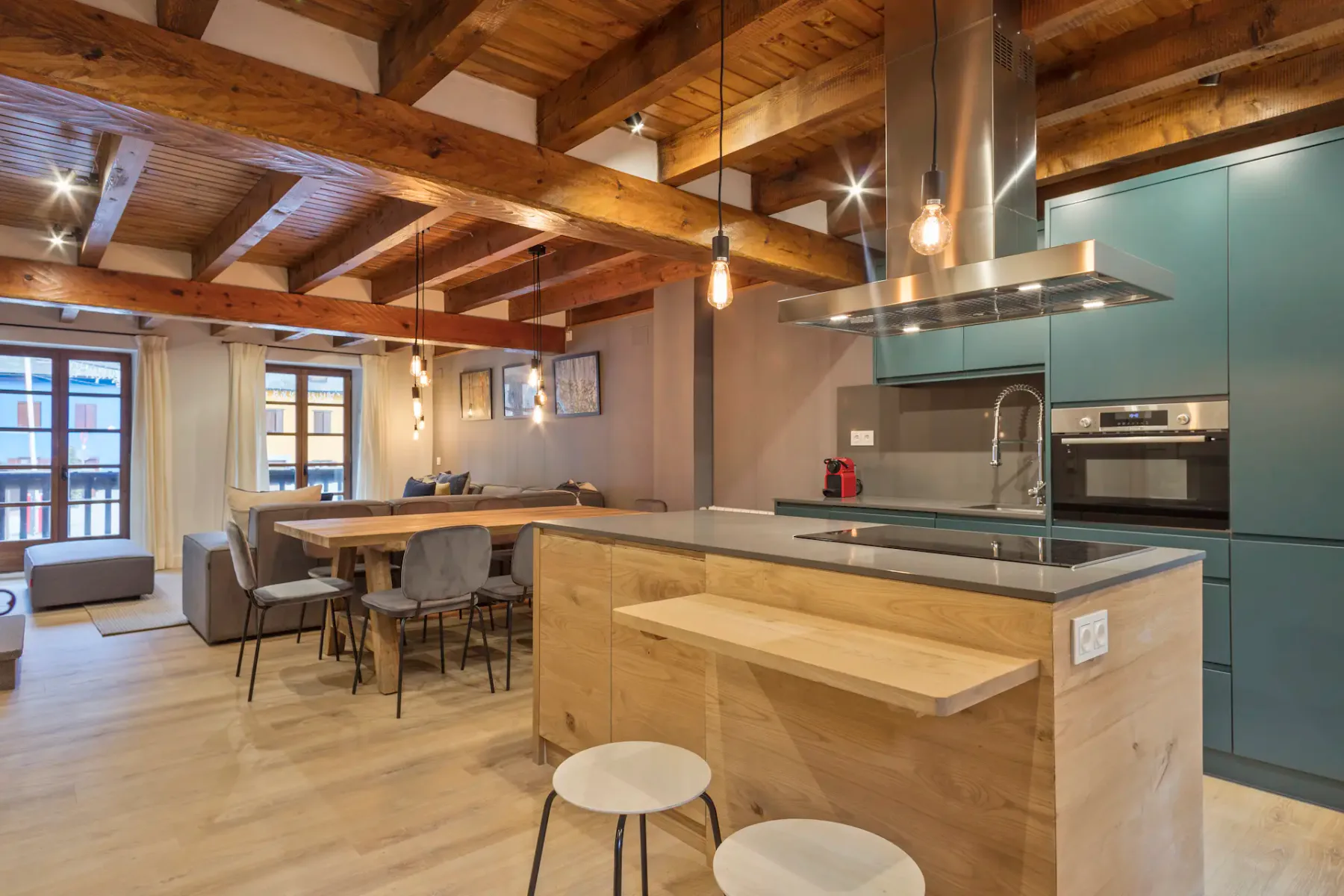 A modern kitchen and dining area with wooden furniture, a table set for four, pendant lights, and large windows allowing natural light. The room features light wood flooring and beige tones.
