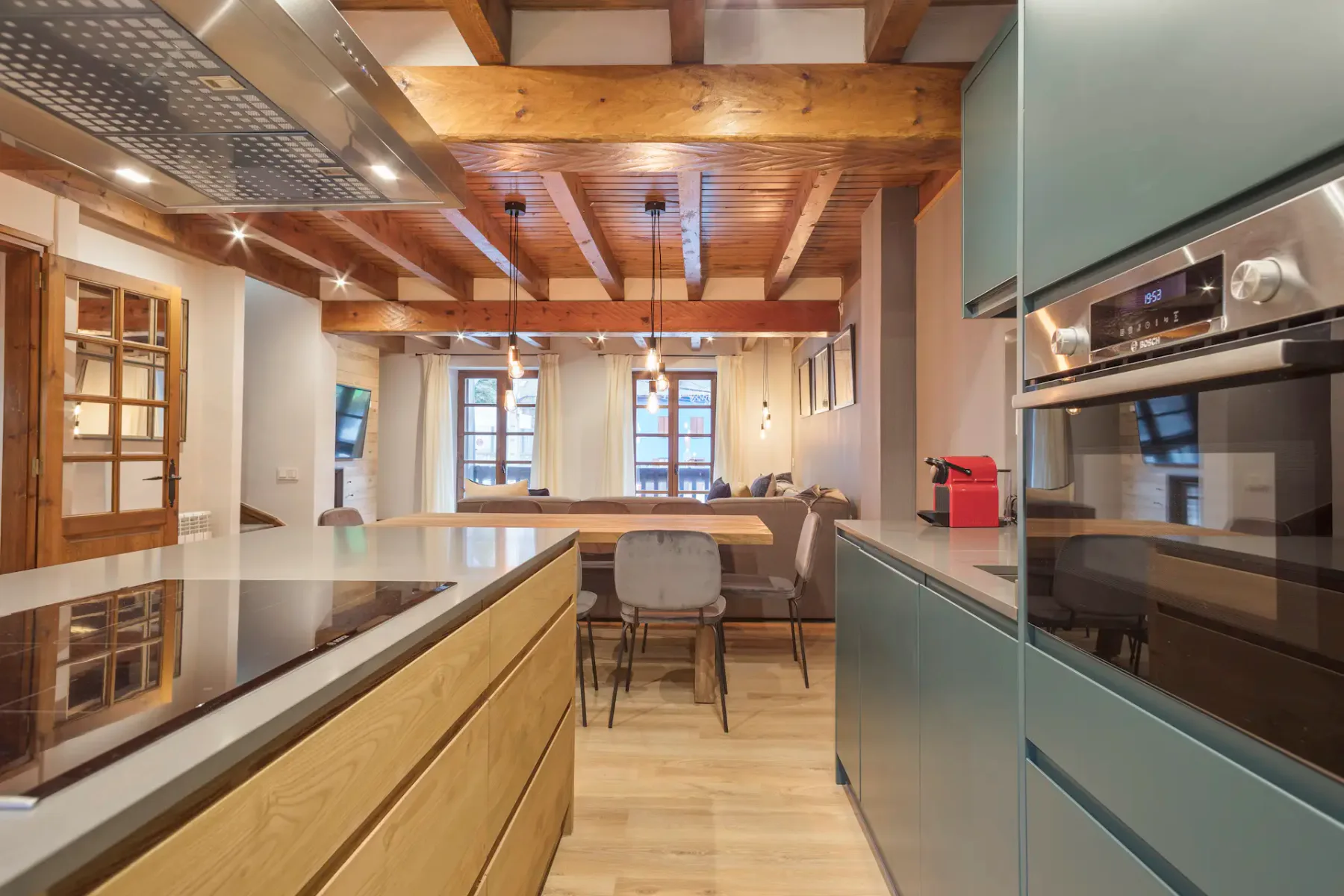 A modern kitchen with a black countertop, light gray cabinets, and wooden flooring. The kitchen opens into a dining area with wooden beams on the ceiling and pendant lights.