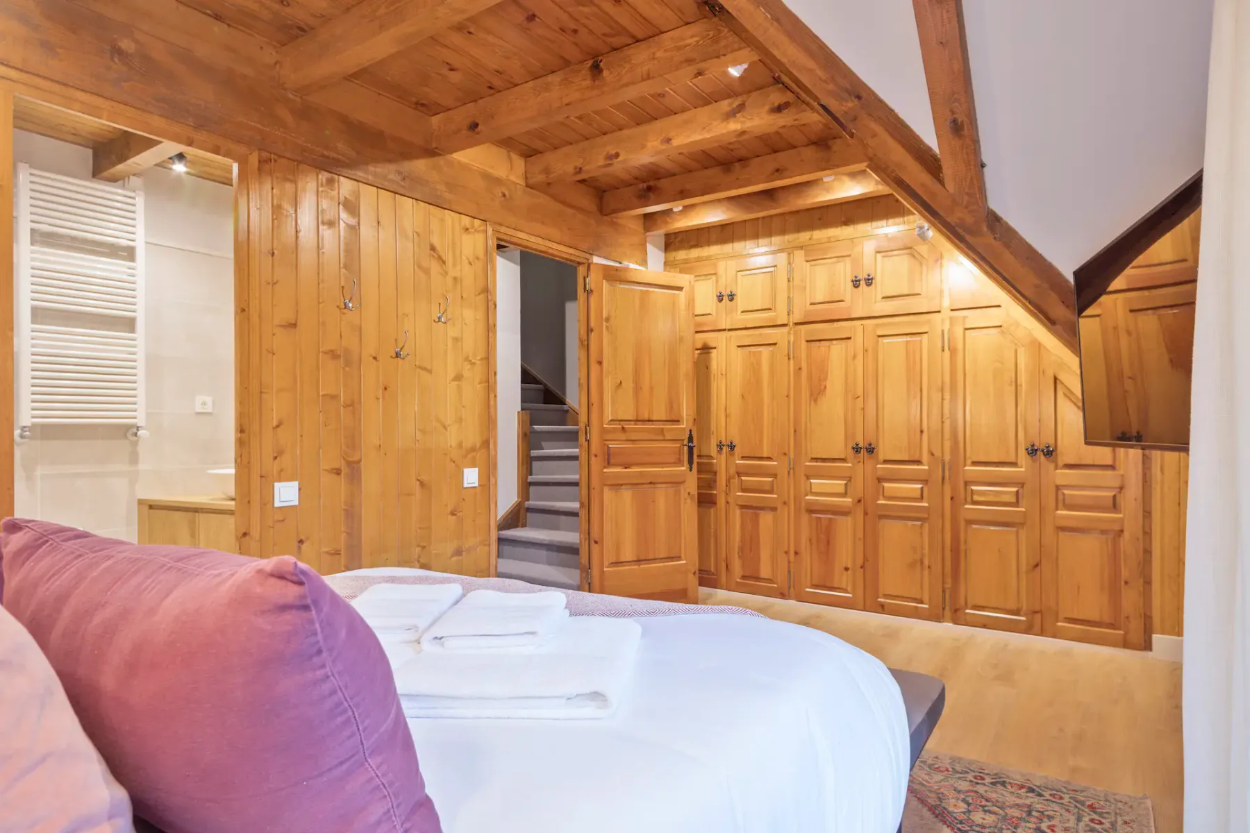 A bedroom with a white bed and purple pillows beneath a wooden ceiling.