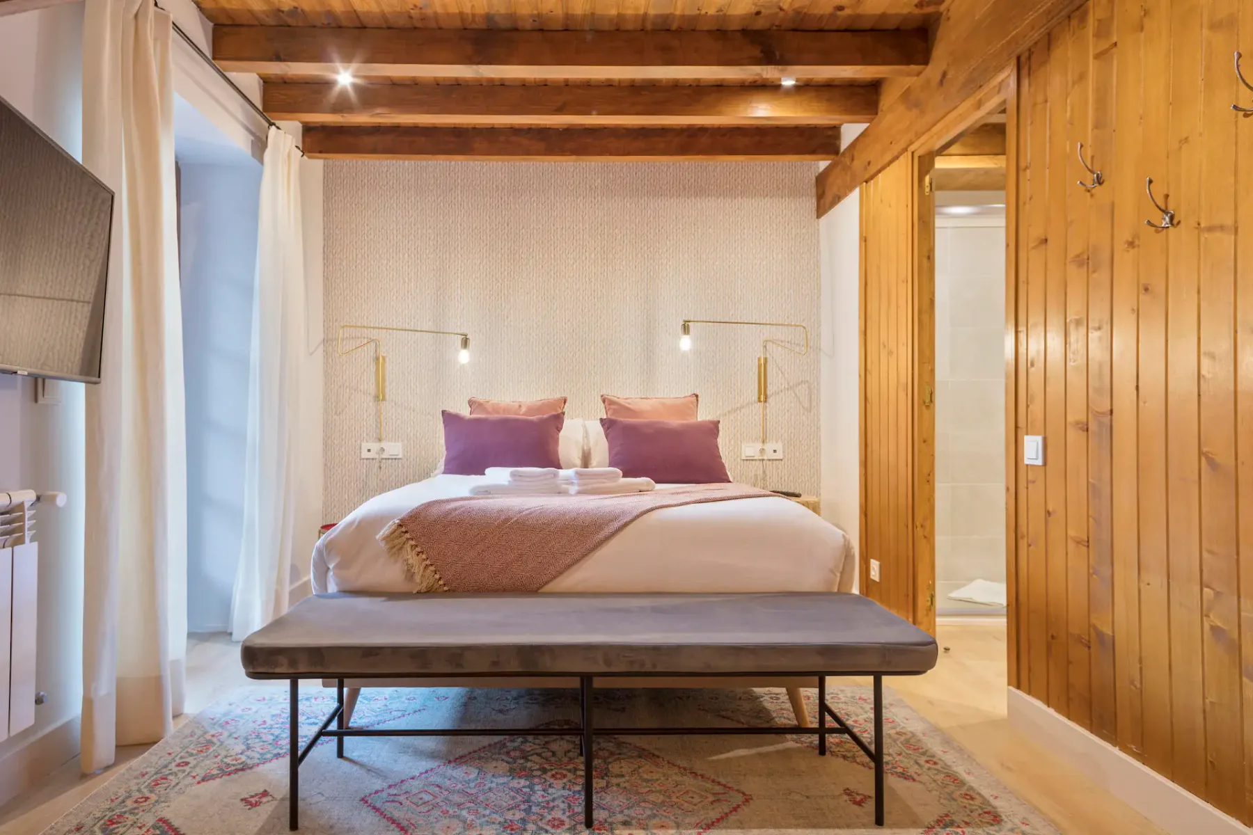 A cozy bedroom with a double bed, purple pillows, a wooden ceiling, and a bench at the foot. A modern bathroom is visible through an open door on the right.