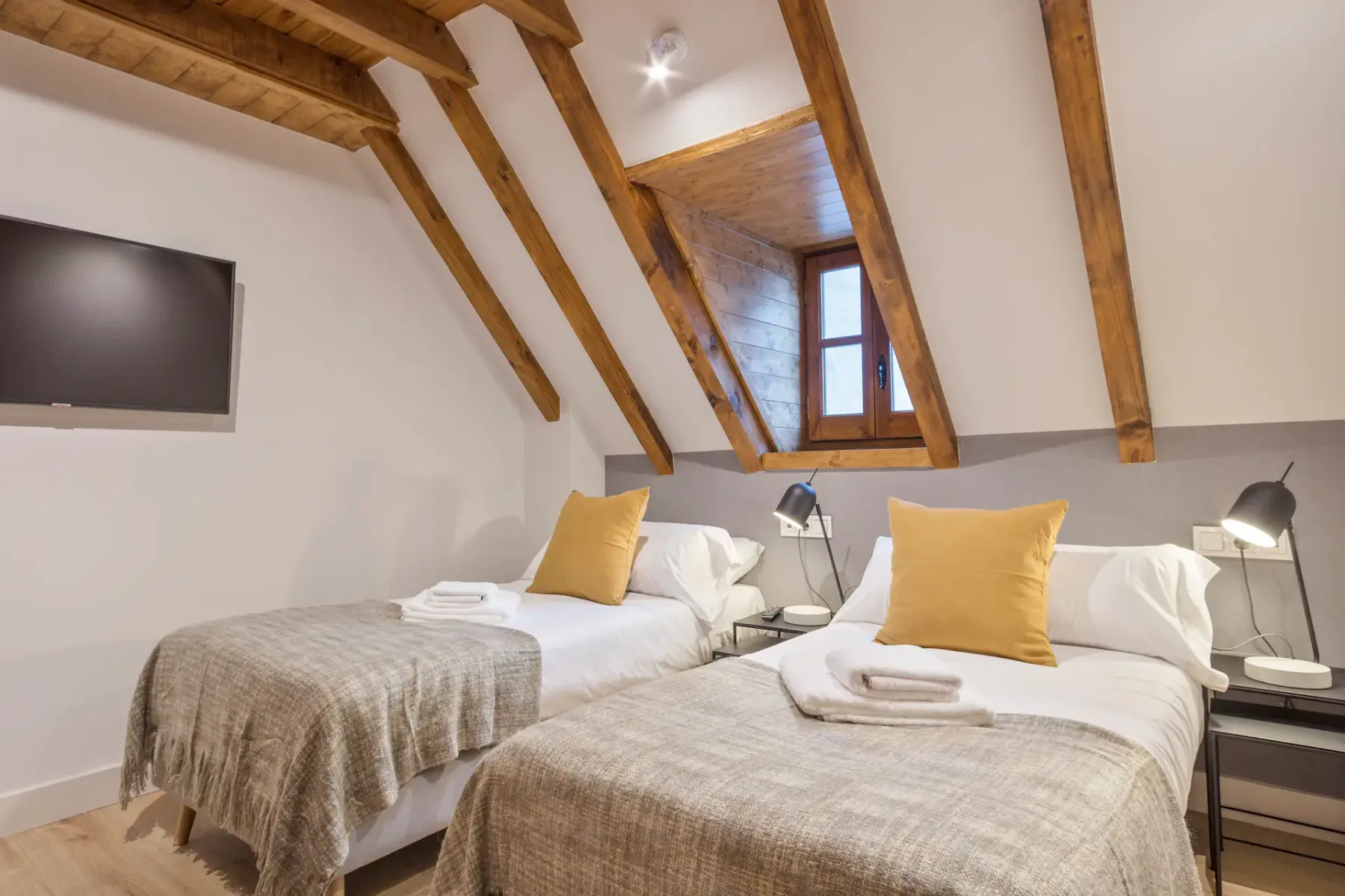 Attic bedroom with two single beds, gray blankets, yellow pillows, a wall-mounted TV, a small window, and wooden beams on the ceiling.