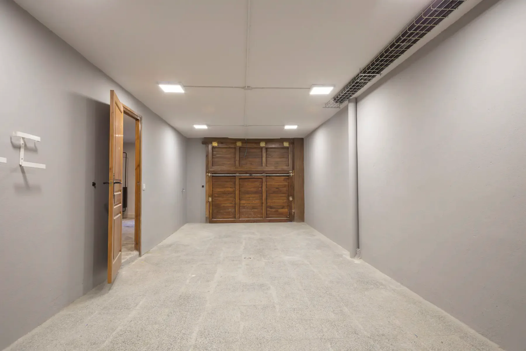 Empty garage with concrete floor, wooden garage door, and side entrance door on the left. Ceiling lights are switched on.
