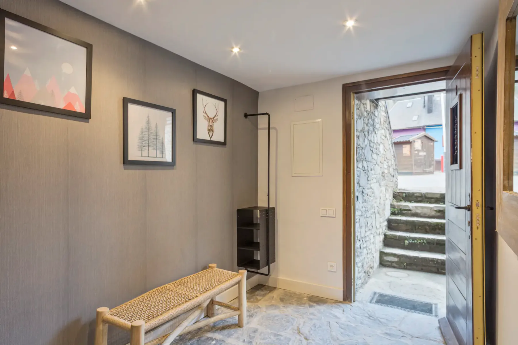 Hallway with a wooden bench, wall art, and a light gray stone floor. The door is open, revealing stone steps leading outside to a street with buildings.
