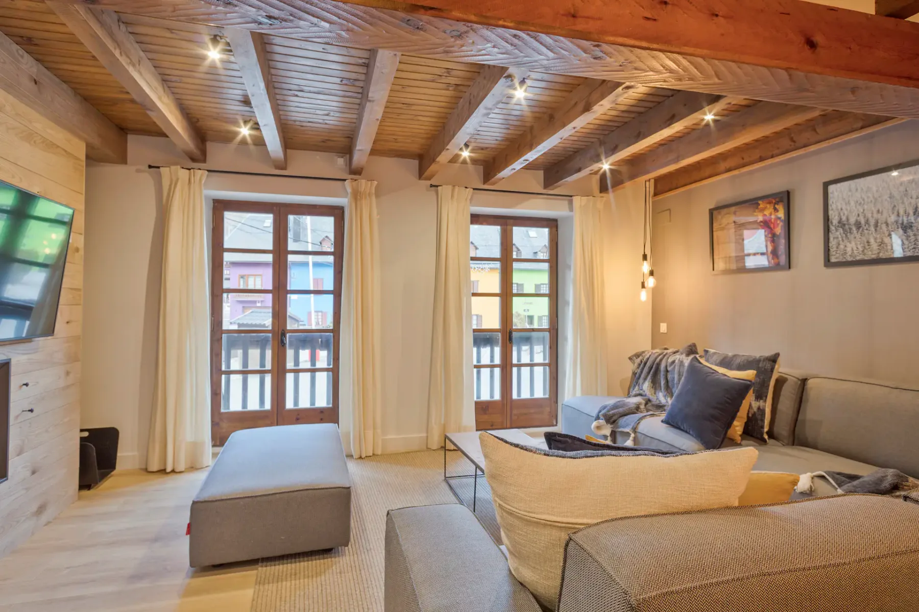 A living room with cushioned seating, an ottoman, and tall windows beneath a wood-beamed ceiling.